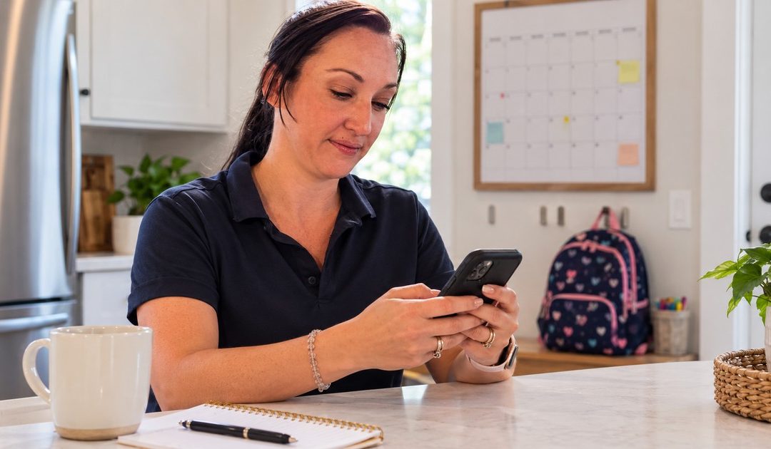 A dark-haired woman drafts a brief co-parenting message on a compact black smartphone held in both hands.